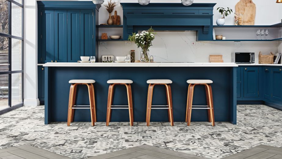 Kitchen space with blue cabinetry and island, with wooden barstools over gray patchwork tile that meets a gray herringbone plank floor