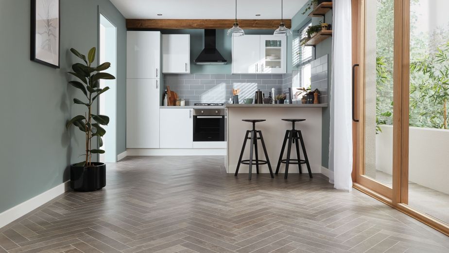 Bright kitchen with gray herringbone tile floor, white cabinetry, small counter with barstools, houseplant, and artwork across from glass doors leading outside