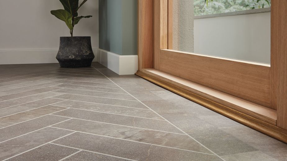 Floor shot highlighting grouted gray herringbone LVT floor and design strip beside wood and glass exterior door, with potted plant in corner