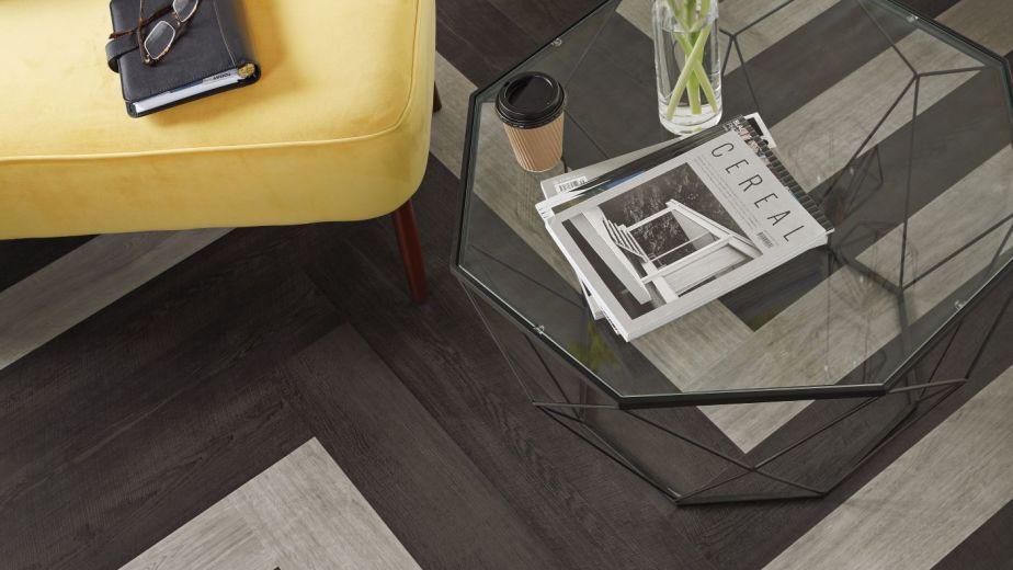 Overhead shot of glass side table, magazines, coffee cup, and yellow gladiolus beside yellow sofa with journal and glasses, with herringbone black and gray plank flooring