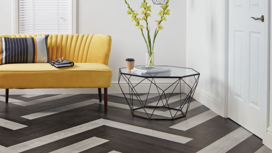 Bright living area featuring bright yellow sofa and glass side table in room with dramatic black and gray herringbone plank floor