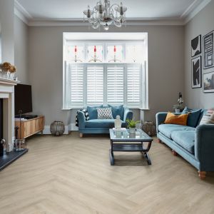 Living room space with taupe herringbone-laid plank floors, stained-glass window behind loveseat, with couch facing coffee table and fireplace topped with vases and hanging artwork