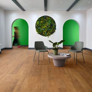 Commercial space with exposed beams on ceiling and brown wood floor, featuring living plant wall decor between two arched doorframes leading to green hallway with blurred woman walking by, with two office chairs by small table in foreground