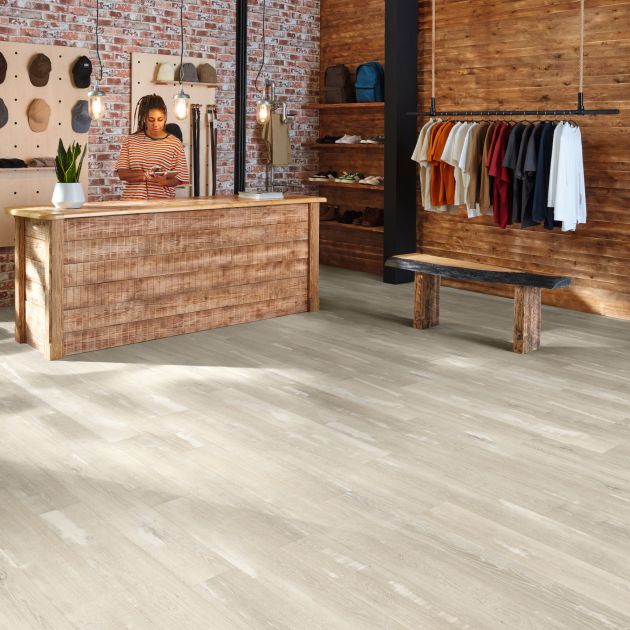 Retail space featuring person behind distressed wooden counter, exposed brick wall, gray patina woodgrain floors, and rack of hanging t-shirts before reclaimed wood bench