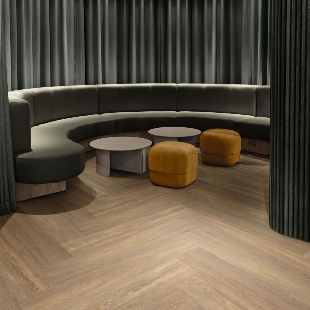 Office space featuring sectional sofa curled around tables and ottoman, partially enclosed by a curtained wall, in space with taupe woodgrain floor installed in a herringbone pattern