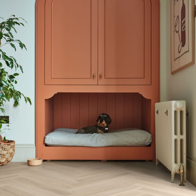 Hallway with pink cabinet, dog bed nook, house plant, radiator, and artwork in room with taupe woodgrain floor installed in herringbone pattern