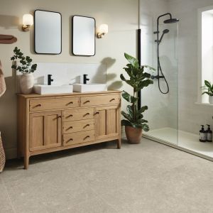 Neutral bathroom with wooden dual sink vanity, mirrors, sconces, towel rack, houseplant, and shower, with taupe square tile floor