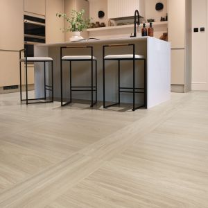Minimalist kitchen with pale woodgrain flooring installed in a grid pattern, with barstools pulled from island and cabinetry and appliances in background