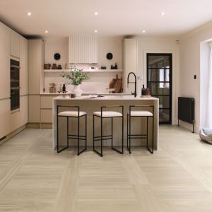 Minimalist kitchen with taupe cabinetry, island with bar stools, houseplant, pet bed by exterior door, and pale woodgrain floor installed in a grid design
