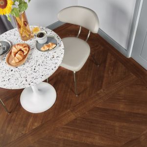Dining space with speckled white table topped with breakfast food, sunflowers in a vase, and two chairs atop reddish-brown plank flooring installed in a V pattern