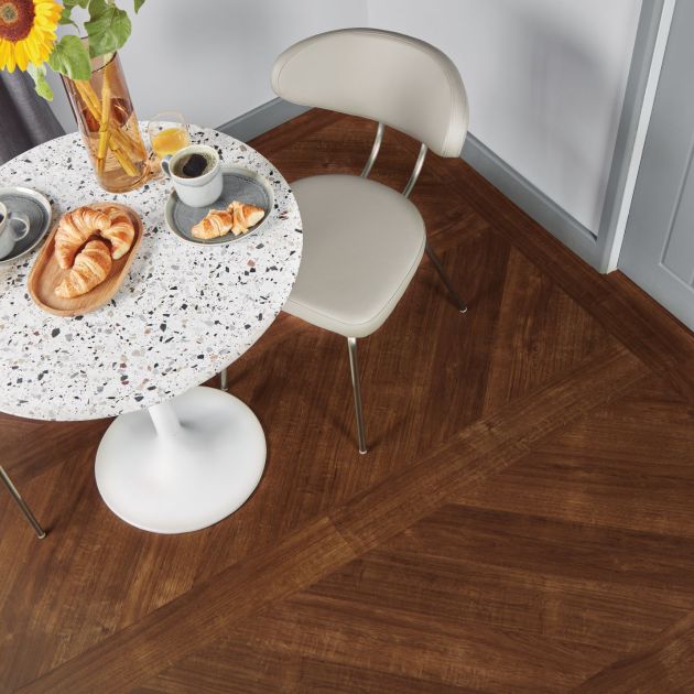 Dining space with speckled white table topped with breakfast food, sunflowers in a vase, and two chairs atop reddish-brown plank flooring installed in a V pattern